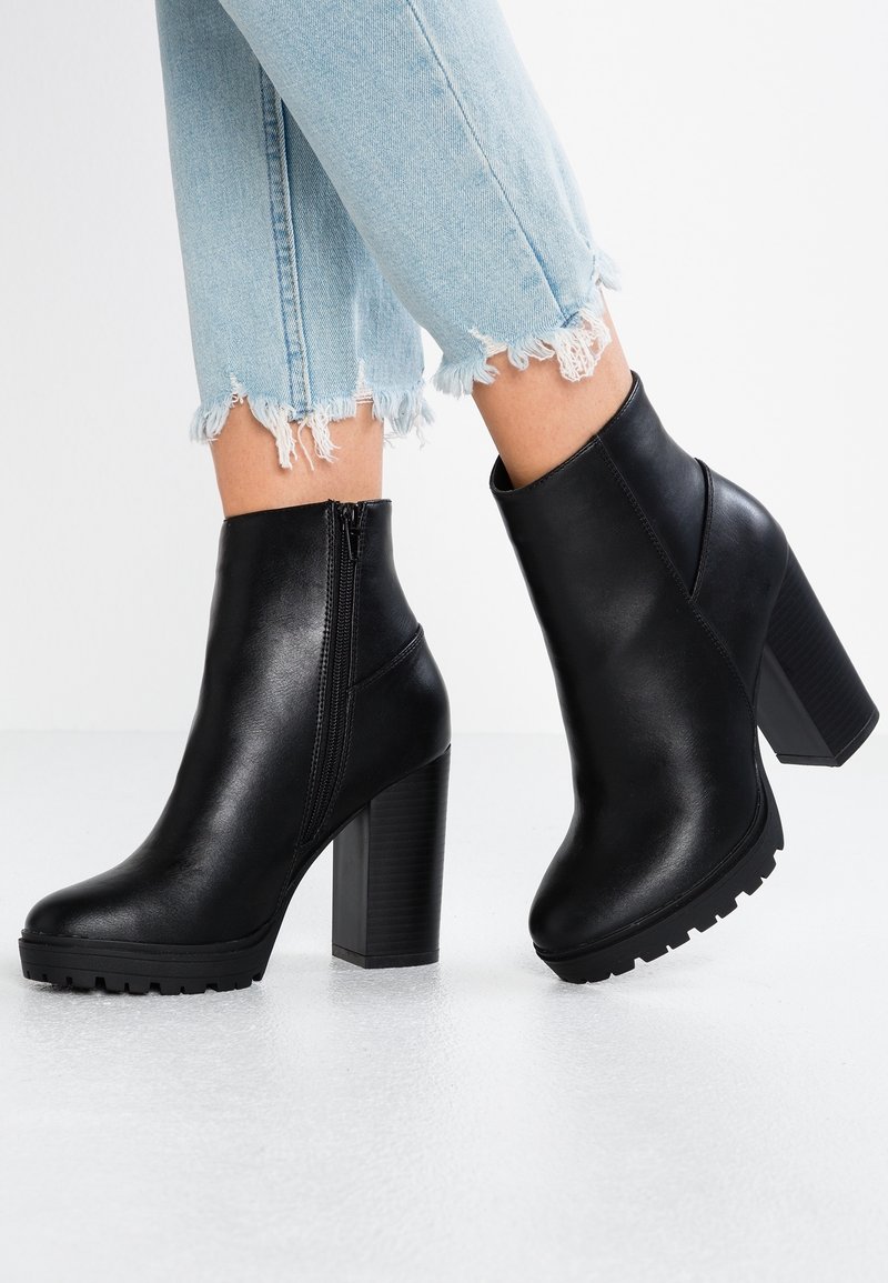 Stivaletti neri in pelle con tacco alto chunky, dettagli con zip laterale e suola testurizzata, abbinati a jeans azzurri sfilacciati.