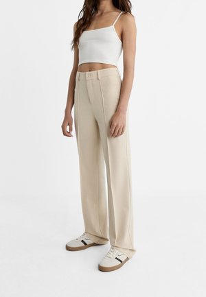 Pantalon beige à jambes larges avec coutures verticales et fermeture par un seul bouton, porté avec un débardeur blanc ajusté et des baskets beiges.