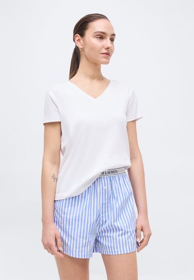 Νεαρή γυναίκα με αλογοουρά, φορώντας λευκή μπλούζα V λαιμόκοψης και μπλε-λευκά κάθετα ριγέ σορτς με ορατό λογότυπο στη μέση.