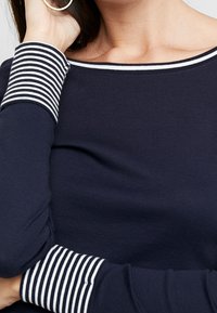 Marineblaues Langarmoberteil mit gestreiften, gerippten Bündchen und Halsausschnitt. Weicher Baumwollstoff mit tailliertem Design, der den Handgelenks- und Schulterbereich betont.