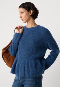 Plavi pleteni pulover s teksturiranim završetkom, okruglim izrezom i peplum rubom. Model drži smeđu zamširanu torbu.
