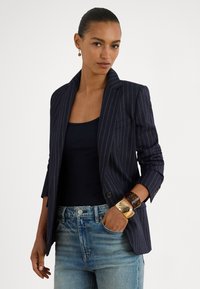 Moteris su atgal sušukuotais plaukais, vilkinti tamsiai mėlyną dryžuotą švarką, juodą marškinėlį be rankovių, mėlynas džinsas, dideli auksiniai ir gyvatės kiauto spalvos apyrankės bei viena auskarą.