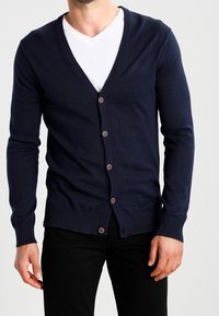 Mann in einer navyblauen Knopfpulloverjacke über einem weißen V-Ausschnitt-Shirt und schwarzen Hosen, der vor einem schlichten Hintergrund steht.