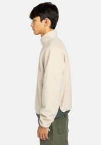 Jasno beżowy polarowy sweter z wysokim kołnierzem i półzamkiem. Posiada luźny krój z kontrastującą granatową dolną częścią i teksturowaną powierzchnią.