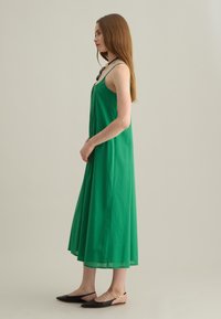 Abito maxi verde senza maniche con una vestibilità ampia. Realizzato in tessuto leggero, presenta una parte anteriore plissettata e una texture liscia; abbinato a scarpe basse nere.