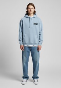 Felpa leggera blu con testo nero "AMOUR", tasca a marsupio, cappuccio con cordino; abbinata a jeans blu chiaro e sneakers bianche.