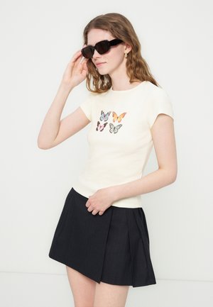 Femeie care poartă ochelari de soare negri, tricou crem cu patru fluturi colorați și fustă plisată neagră, pozând pe un fundal alb.