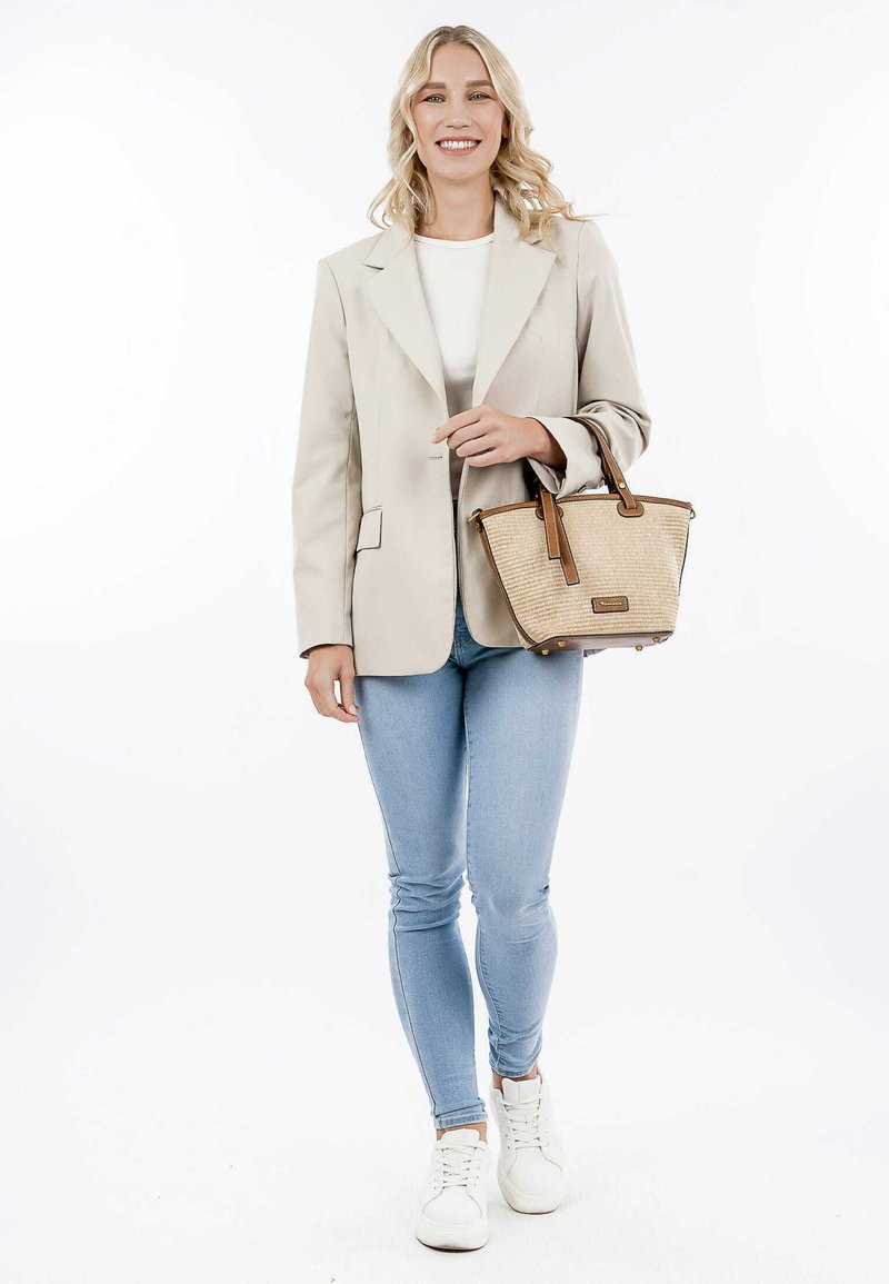 Blazer beige sopra una maglia bianca, abbinato a jeans azzurri chiari e sneakers bianche. La modella tiene una borsa a tracolla intrecciata color marrone chiaro con dettagli marroni.