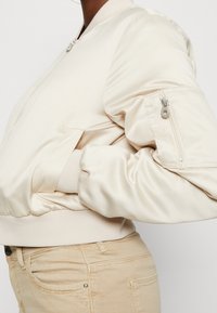 Beżowa satynowa kurtka bomberka z zamkiem błyskawicznym na rękawie i ściągaczami, noszona z jasnobeżowymi dżinsami, ręka włożona w kieszeń kurtki.