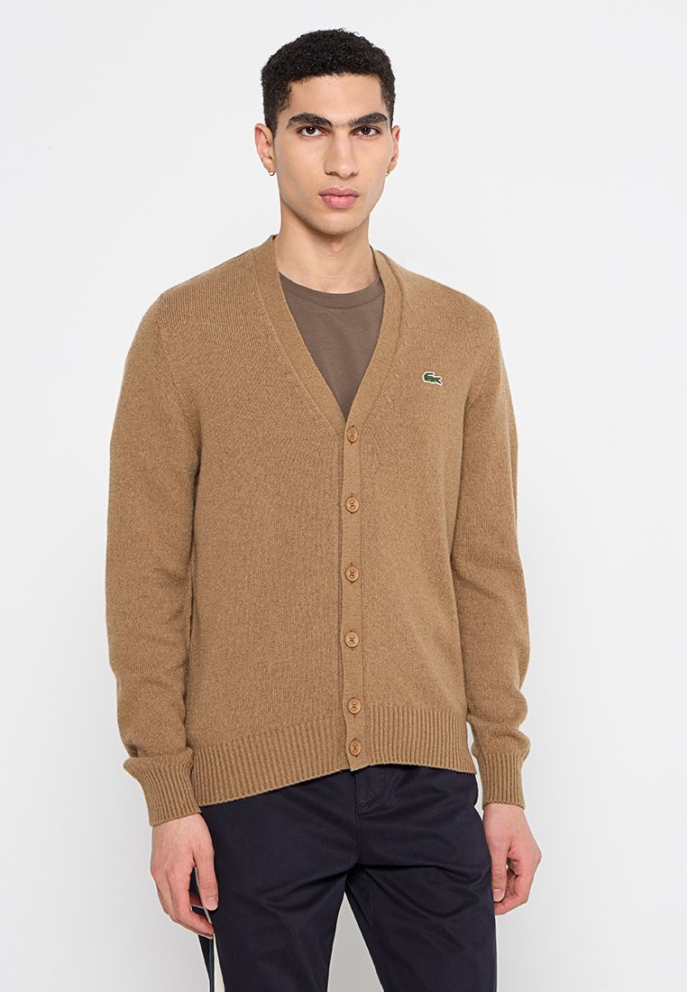 Cardigan en laine beige avec un col en V, des poignets côtelés et cinq boutons marron. Présente un petit logo Lacoste vert sur la poitrine gauche.