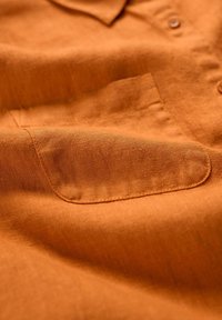 Hedvábná košile s krátkým rukávem v oranžové barvě, s měkkou texturou, která má náprsní kapsu a knoflíky. Tkanina vypadá lehká s jemným vzorem tkaní.
