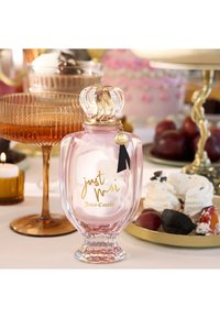Flacon de parfum Juicy Couture rose avec bouchon doré en forme de couronne posé sur une table, entouré d'assiettes à dessert, de fruits, d'une bougie et d'un verre.