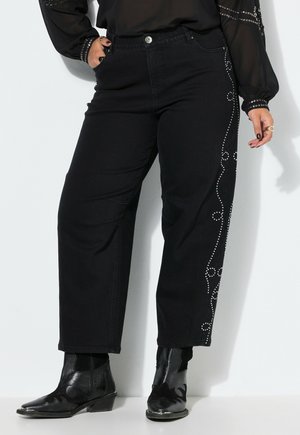 Persoon met een zwarte broek met decoratieve studs aan de zijkanten, een zwarte doorschijnende blouse en zwarte enkellaarsjes, staand tegen een witte muur.
