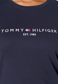 T-shirt in cotone blu navy con testo ricamato bianco "TOMMY HILFIGER" e "EST. 1985" con un piccolo logo in rosso e blu. Texture liscia.