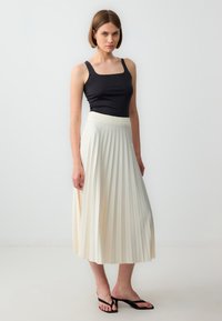 Zwarte aansluitende tanktop en crèmekleurige geplooide midi-rok met elastische tailleband, gecombineerd met zwarte flip-flops op een neutrale achtergrond.