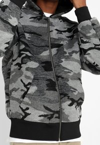 Sudadera con cremallera de camuflaje gris con una textura suave, acentos negros, capucha y bolsillos frontales. Presenta tonos variados de gris y negro.