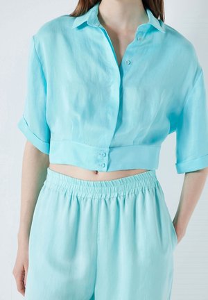 Chemise légère bleue à manches courtes, à coupe courte, avec un col et deux boutons à l'avant, assortie à un pantalon à taille élastique.