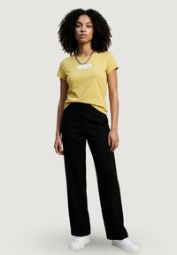 Mujer joven con cabello rizado, vistiendo una camiseta amarilla, pantalones negros, zapatillas blancas y un collar de cadena plateada, de pie sobre un fondo liso.
