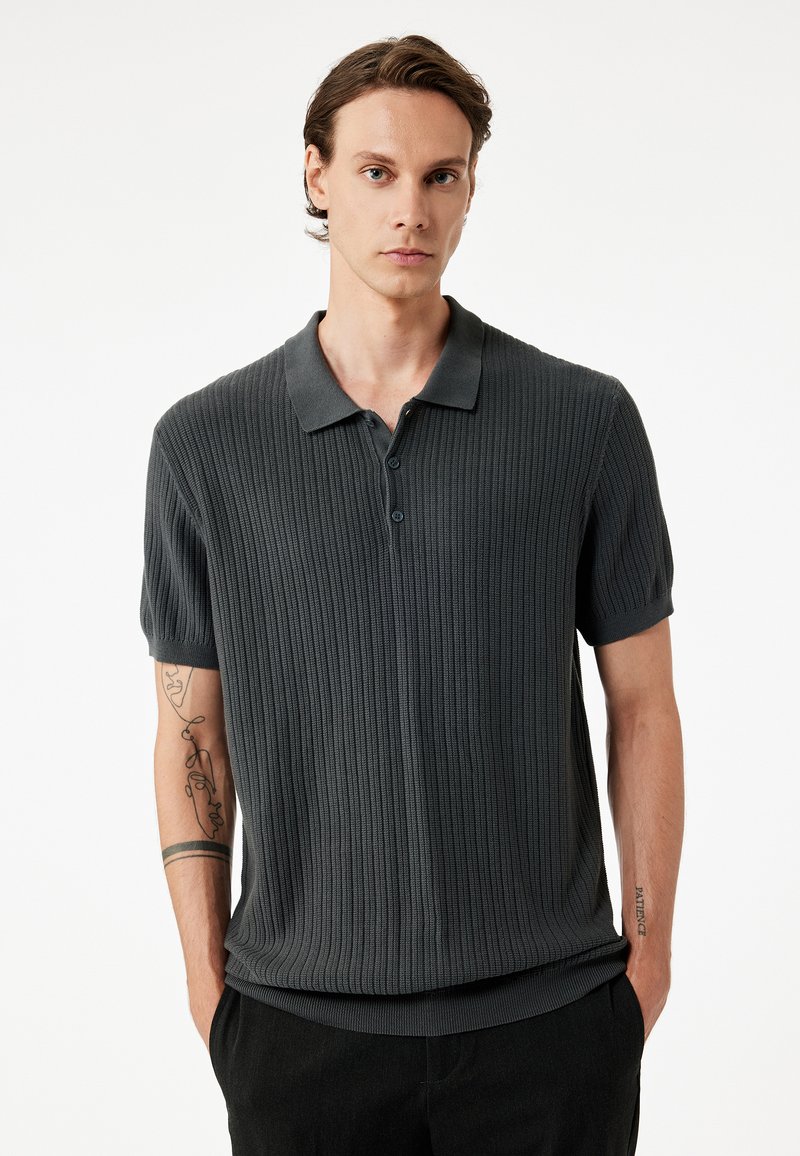 Donkergrijze polo shirt met korte mouwen en ribbeltextuur, drie-knopen sluiting en contrasterende zachte kraag. Gedragen met de handen in de zakken.