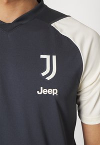 Camiseta deportiva de un azul marino oscuro con mangas cortas color crema. Presenta un logo con dos rayas blancas y "Jeep" en blanco debajo. Textura suave.