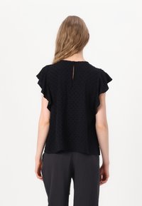 Blouse noire avec des manches volantes et une texture à pois, dotée d'un détail en ouverture dans le dos. Le tissu est léger, créant une apparence fluide.