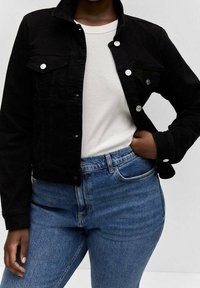 Giacca di denim nera con colletto, chiusura frontale con bottoni, due tasche sul petto e maniche lunghe, abbinata a una camicia bianca e jeans blu.