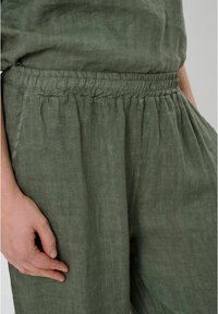 Zelene lanene hlače z elastičnim pasom, opremljene s pliči in stranskimi žepi. Material je nekoliko teksturiran in mehak na otip.