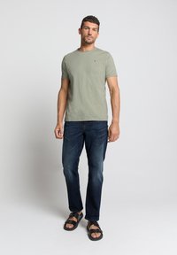 Olijfgroene t-shirt met korte mouwen, gecombineerd met donkerblauwe jeans en zwarte sandalen. De t-shirt heeft een klein logo op de borst.