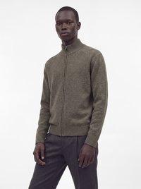 Homme portant un pull zippé marron avec col et poignets côtelés, et un pantalon gris foncé, debout devant un fond blanc uni.