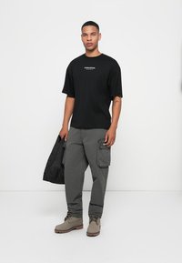 Jonge man in een zwart "Studio Edition" T-shirt, grijze cargobroek, beige laarzen, met een zwarte jas op een witte achtergrond.