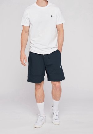 Hombre que lleva puesta una camiseta blanca de manga corta, pantalones cortos azul marino, calcetas blancas y zapatillas blancas, de pie contra un fondo liso.