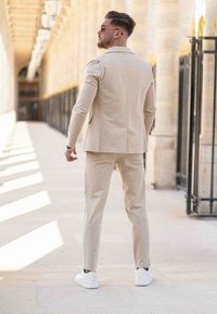 Costume beige sur mesure avec des manches longues et des jambes droites, associé à des baskets blanches. Tissu lisse et design structuré.