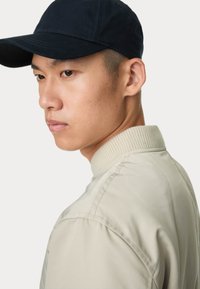 Beige bomberjack met geribbelde kraag, gecombineerd met een marineblauwe baseballpet. Gladde stofstructuur, zichtbare stikdetails op de jas.