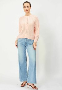 Vaaleanpunainen neule, jossa on pitsikuvio, pyöreä kaula-aukko ja napit. Yhdistetty leveälahkeisiin haalistuneisiin sinisiin farkkuihin ja ruskeisiin korkokenkiin.