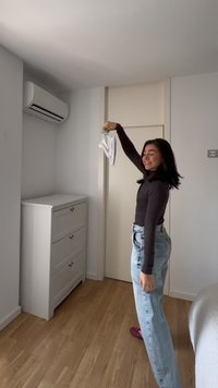 Jeune femme souriante, tenant un tissu blanc ou un vêtement au-dessus de sa tête dans une pièce minimaliste avec un sol en bois et une commode blanche.
