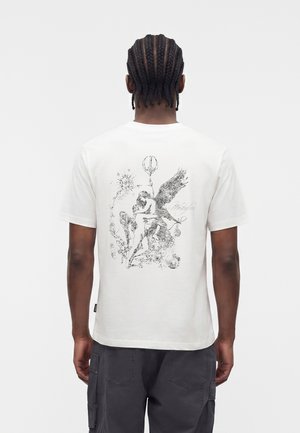 Άνδρας με πλεξούδες που φοράει λευκό μπλουζάκι με λεπτομερή μαύρο σκίτσο με φτερωτές και ανθρώπινες φιγούρες στην πλάτη.