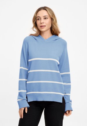 Frau trägt einen hellblauen Hoodie mit weißen horizontalen Streifen und schwarzen Hosen, steht vor einem schlichten weißen Hintergrund.