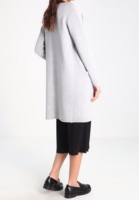 Grå, ribbade lång cardigan med sidofickor, matchad med en svart knälång kjol och svarta skor, sedd bakifrån.