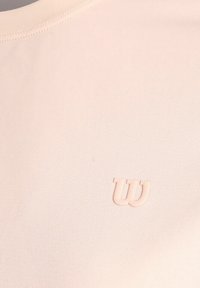 Lichtroze stof met een verhoogd, zacht "W" logo dat op de borst geïmprint is nabij de halslijn.