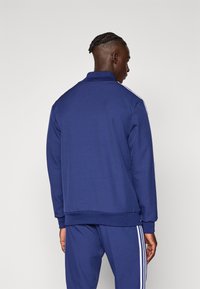 Σκούρο μπλε αθλητικό μπουφάν με ψηλό γιακά, ριμπ μανσέτες και τελείωμα. Διακοσμημένο με λευκές ρίγες στους ώμους. Μαλακό, υφασμάτινο με υφή.