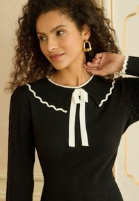 Fekete kötött felső fehér hullámos szegéllyel rendelkező gallérral, fodros nyakkendővel és texturált ujjak.