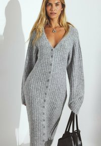 Gray ribbed knit cardigan dress with a deep V-neck, long sleeves, and buttons down the front. Carries a black tote bag.