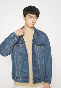 Chaqueta de denim en azul medio con ligeras desgastes, cierre de botones, dos bolsillos frontales y costuras en contraste sobre una camisa beige.