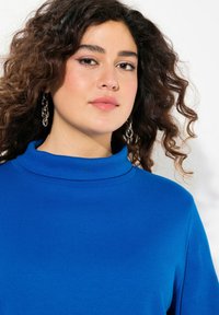 Maglione a collo alto blu a costine con maniche lunghe. Texture morbida e vestibilità comoda. Modella che indossa orecchini a catena argentati su uno sfondo chiaro.
