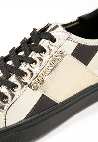 Gros plan sur une basket Arman Jeans noire et beige avec des lacets noirs et une plaque métallique avec logo sur le côté.