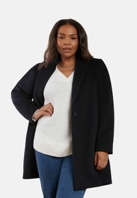 Svart yllekappa med slagkrage, enkel knappstängning och sidofickor, bärs över en vit stickad tröja och blå jeans.