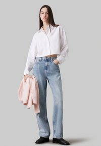 Camisa blanca de botones cortada, jeans rectos azul claro, sosteniendo un suéter rosa suave. Botines negros completan el atuendo.