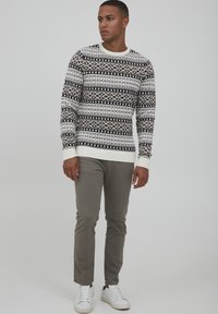Pull en tricot blanc avec des motifs géométriques noirs, poignets et col côtelés ; associé à un pantalon slim gris clair et des baskets blanches.