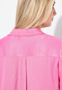 Pink skjorte med en glat tekstur, der har en klassisk krave og en plissé detalje bagpå. Stoffet virker let og let gennemsigtigt.