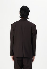 Ruskea räätälöity bleiseri, jossa on selkeä muotoilu, yksittäinen halkio, pitkät hihat ja hienovaraiset napin yksityiskohdat hihoissa. Sujuva, muodollinen kangasrakenteeltaan.